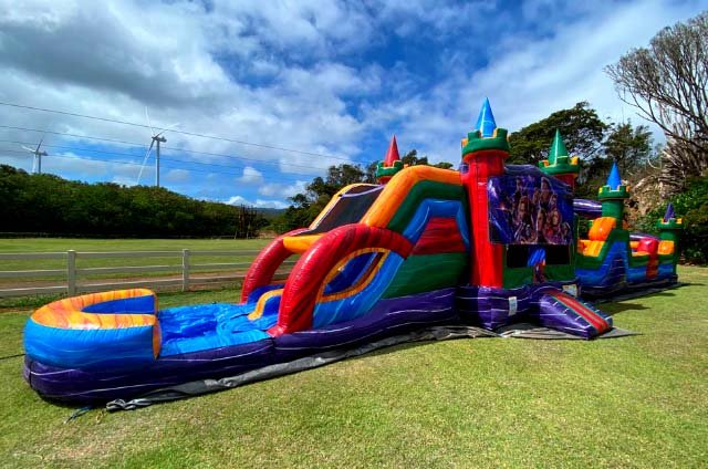 A large rainbow combo bounce house with a slide set up on grass at an Oahu park
