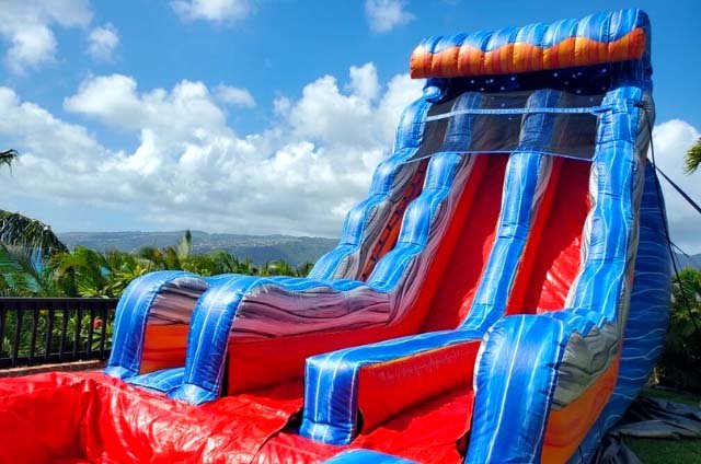 A tall blue and red inflatable water slide set up outdoors with the Oahu mountains and a bright sky behind it