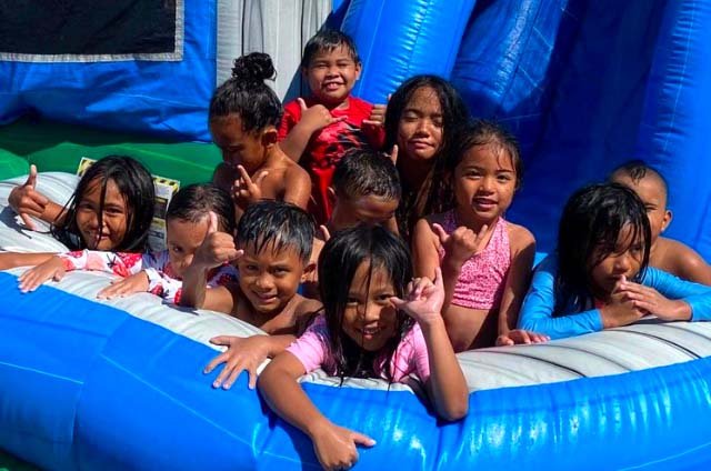 A group of kids piled together in an inflatable slide pool, soaked and throwing shakas at an Oahu party