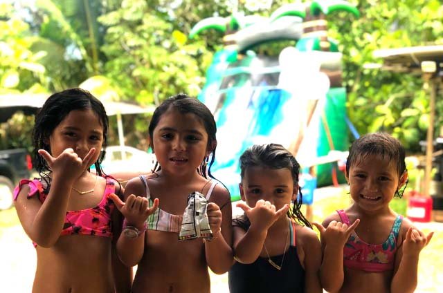 Four kids in swimsuits throwing shakas in front of a green inflatable water slide